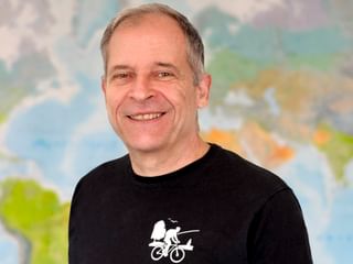 Portrait von Christoph mit kurzem grauen Haar, der breit lächelt. Er trägt ein schwarzes Shirt mit weißer Fahrradgrafik. Eine bunte Karte ist hinter ihm sichtbar.