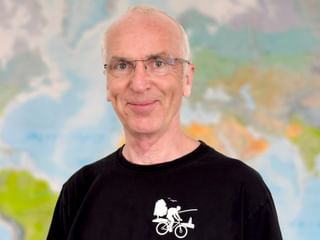 Portrait von Dieter mit Brille und schwarzem Radfahrer-Shirt mit weißer Radfahrer-Grafik, stehend vor einer bunten Weltkarte.
