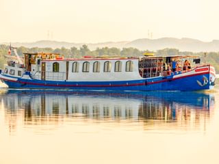 MS L'Estello, ein blau-weißes Flusskreuzfahrtschiff mit Bogenfenstern, gleitet auf ruhigem Wasser mit sichtbarer Spiegelung. Grünes Ufer im Hintergrund.