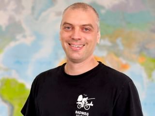 Portrait von Eugen mit kurzen blonden Haaren und blauen Augen, lächelnd in einem schwarzen Radweg-Reisen T-Shirt. Eine bunte Weltkarte ist im Hintergrund sichtbar.