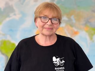 Suzanne mit Brille und schwarzem Radweg-Reisen T-Shirt mit Radfahrer-Logo, stehend vor einer Weltkarte im Hintergrund.