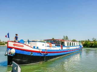 MS Caprice, ein weiß-blaues Flusskreuzfahrtschiff mit rotem Rand, liegt an einem ruhigen Wasserweg. Eine Person steht an Deck unter blauem Himmel.