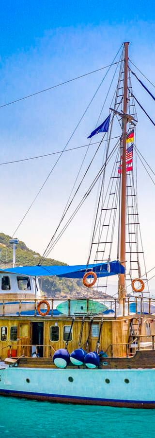MS Panagiota, ein traditionelles Segelschiff mit blau-weißem Rumpf, vor Anker in türkisfarbenem Wasser bei Parga mit grünen Hügeln im Hintergrund.