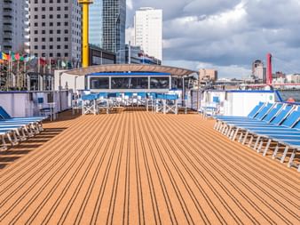 Geräumiges Sonnendeck der MS Olympia mit Reihen blauer Liegen auf Holzplanken. Internationale Flaggen und Stadtskyline im Hintergrund.