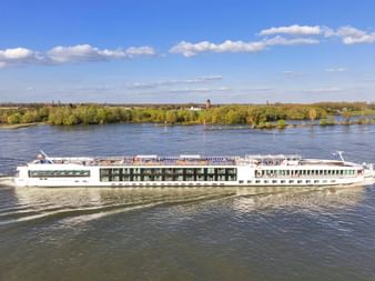 Weißes Flusskreuzfahrtschiff MS Lisabelle auf der Donau mit grünen bewaldeten Ufern und blauem Himmel mit weißen Wolken im Hintergrund.