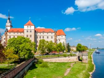 Schloss Hartenfels in Torgau mit orangefarbenen Dächern und Turm, umgeben von grünen Bäumen entlang der Elbe unter blauem Himmel.