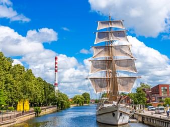 Großes Segelschiff mit weißen Segeln liegt im Kanal von Klaipeda, Litauen. Grüne Bäume säumen den Wasserweg unter blauem Himmel mit weißen Wolken.