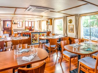 Heller Speisesalon auf der MS Caprice mit Holztischen, gedeckt mit Gläsern und Servietten, gepolsterten Stühlen, großen Fenstern mit Vorhängen und Barbereich.