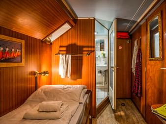 Kompakte Kabine auf MS L'Estello mit holzgetäfelten Wänden, Einzelbett mit weißer Bettwäsche, Wandlampe und eigenem Bad durch offene Tür sichtbar.