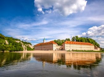 Kloster Weltenburg mit orangefarbenen Ziegeldächern spiegelt sich in der ruhigen Donau, umgeben von grünen bewaldeten Klippen unter blauem Himmel.