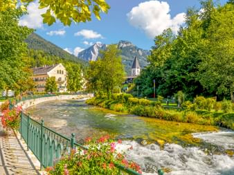 Fluss Traun mit Stromschnellen fließt durch Bad Aussee, gesäumt von blumengeschmückten Geländern. Kirchturm und Berge unter blauem Himmel.