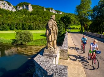 Zwei Radfahrer überqueren eine Steinbrücke mit Statue bei Hausen im Tal. Weiße Kalksteinfelsen und grüne Wälder säumen das Donautal unter blauem Himmel.