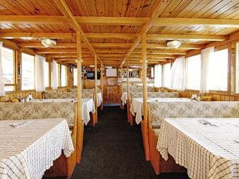 Innenraum des MS Aneta Salons mit Holzdeckenbalken, Bänken mit Blumenmuster, karierten Tischdecken und großen Fenstern mit weißen Vorhängen.
