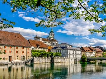 Historische Gebäude entlang der Aare in Solothurn mit Kuppel und Turm der St. Ursen-Kathedrale im Hintergrund, eingerahmt von grünen Ästen.