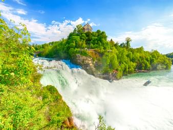 Rheinfall bei Schaffhausen mit kraftvoll herabstürzendem Wasser, üppiger grüner Vegetation und einem historischen Gebäude auf dem Hügel.