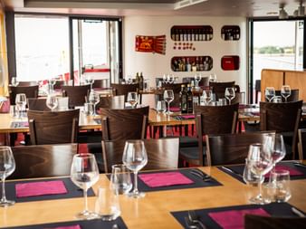 Helles Restaurant-Interieur mit Holztischen, Weingläsern und rosa Servietten. Wandregale zeigen Weinflaschen. Große Fenster mit Wasserblick.