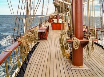 Holzdeck des Großseglers Atlantis mit Mast, Takelage und aufgerollten Seilen. Das Schiff segelt auf blauem Meer unter teils bewölktem Himmel.