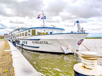 Weißes Flusskreuzfahrtschiff MS Rigoletto mit blauem Rand am Betonpier vertäut. Das Schiff hat große Fenster und Flaggen auf dem Oberdeck.