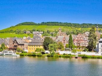 Traben-Trarbach an der Mosel mit Fachwerkhäusern, einem Flussschiff und weinbergbedeckten Hügeln unter blauem Himmel.