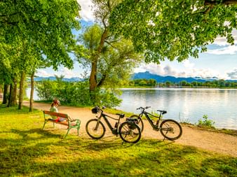 Radfahrer entspannt auf einer Bank am Chiemsee mit zwei abgestellten Fahrrädern. Berge und grüne Bäume rahmen die malerische Seelandschaft.