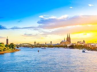 Der Kölner Dom mit seinen Zwillingstürmen dominiert die Skyline am Rhein bei Sonnenuntergang. Eine Brücke und Boote sind auf dem Wasser unter goldenem Himmel sichtbar.