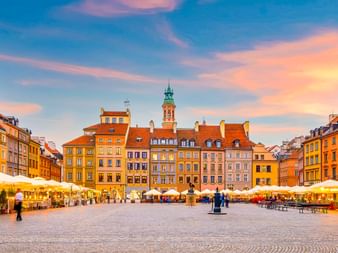 Warschauer Altstadtmarkt mit bunten historischen Gebäuden, Straßencafés und Kopfsteinpflaster unter blauem und orangefarbenem Abendhimmel.