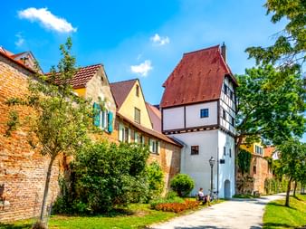Weißer mittelalterlicher Torturm mit Fachwerkaufbau und rotem Dach in Donauwörth, umgeben von historischen Gebäuden und grünen Bäumen.