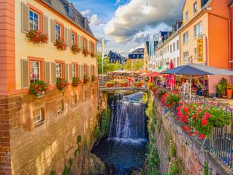 Wasserfall in Saarburg zwischen bunten historischen Gebäuden mit Blumenkästen. Straßencafé mit Sonnenschirmen auf der rechten Seite.