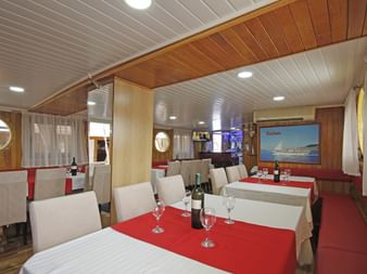 Innenraum des MS Kazimir Speiseraums mit weiß gedeckten Tischen, roten Läufern, Weingläsern und einem Bildschirm mit dem Schiff. Holzverkleidung und Bullaugen.