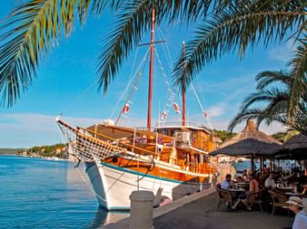 Traditionelles Holzsegelschiff MS Orkan mit weiß-blauem Rumpf an einer Uferpromenade vertäut. Palmen rahmen die Szene, eine Café-Terrasse ist sichtbar.