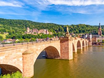 Heidelberger Alte Brücke mit roten Sandsteinbögen über dem Neckar, mit Heidelberger Schloss am bewaldeten Hang und Stadtgebäuden.