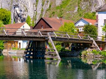 Radfahrer überqueren eine historische Holzbrücke über die Altmühl in Essing, mit weißem Turm und Kalksteinfelsen im Hintergrund.