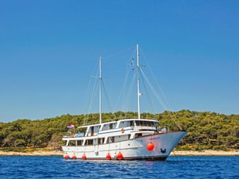 Weiße Motoryacht MS Azimut mit zwei Masten und roten Fendern vor Anker auf ruhigem blauen Wasser, mit grüner bewaldeter Küste im Hintergrund.