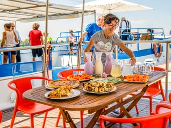 Crew-Mitglied serviert Essen und Getränke auf dem Sonnendeck der MS Caprice. Tisch mit Gebäck, Obst und Getränken. Passagiere entspannen auf blau-weißem Bootsdeck.