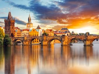 Karlsbrücke über die Moldau in Prag bei Sonnenuntergang, mit historischen Steinbögen, die sich im ruhigen Wasser spiegeln und Altstadtsilhouette.