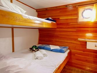 Holzkabine auf der MS Mirabela mit Etagenbetten, weißer Bettwäsche, blauen Handtüchern und rundem Bullauge mit gelben Vorhängen.