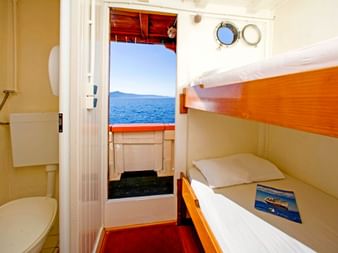 Kompakte Kabine auf der MS Dalmatino mit Holz-Etagenbetten, weißer Bettwäsche und eigenem Bad. Offene Tür zeigt Deck mit Meerblick und Bullaugen.
