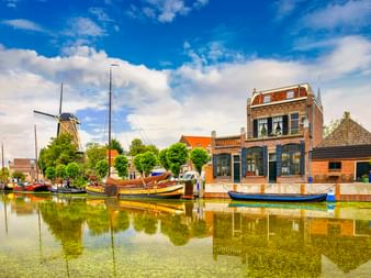 Kanal in Gouda mit traditionellen Booten, historischen Backsteingebäuden und einer Windmühle unter blauem Himmel mit weißen Wolken.