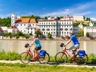 Zwei Radfahrer auf roten Rädern mit blauen Taschen fahren entlang der Donau in Passau. Historische Gebäude mit orangen Dächern am Flussufer.