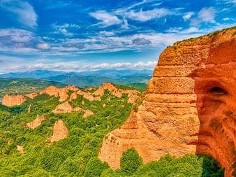 Dramatische orange-rote Felsformationen inmitten grüner Vegetation im Naturpark Las Médulas, Spanien, mit Bergen im Hintergrund.
