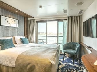 Moderne Kabine auf der MS Swiss Crown mit Doppelbett, türkisfarbenen Kissen, Sessel, TV und großen Fenstern mit Flussblick.