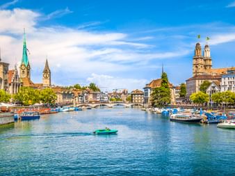 Die Limmat fließt durch Zürich mit Booten am Ufer, historischen Kirchtürmen und dem Grossmünster unter blauem Himmel.