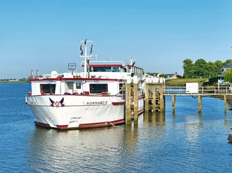 Weißes Passagierschiff MS Normandie mit rotem Zierstreifen an einem Holzsteg vertäut. Blaues Wasser und klarer Himmel sichtbar, mit grünen Bäumen am Ufer.