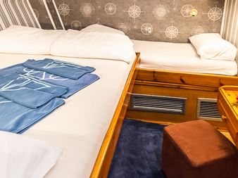 Kabineninterieur auf der MS Panagiota mit Doppelbett, weißer Bettwäsche, blauen Handtüchern, Holzmöbeln und braunem Hocker am Fußende.