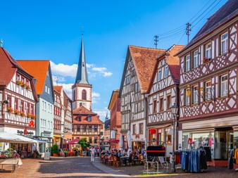 Bunte Fachwerkhäuser säumen eine Fußgängerzone in der Lohrer Altstadt. Ein Kirchturm erhebt sich im Hintergrund unter blauem Himmel.