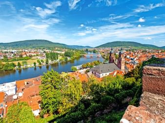 Luftaufnahme von Miltenberg mit roten Ziegeldächern, Main und grünen Hügeln. Kirchturm zwischen historischen Gebäuden unter blauem Himmel.