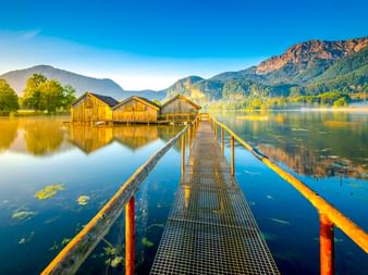Metallsteg führt zu drei Holzbootshäusern am ruhigen Kochelsee. Berge und Bäume spiegeln sich im Wasser unter blauem Morgenhimmel.