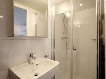 Modernes Badezimmer auf der MS Magnifique II mit weißem Waschbecken, Spiegelschrank und verglaster Dusche mit Handbrause.