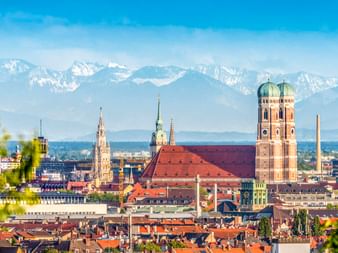 Münchner Stadtbild mit den Zwillingstürmen der Frauenkirche und rotem Dach. Schneebedeckte Alpen im Hintergrund unter blauem Himmel.