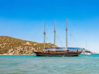 Dreimastiges Holzsegelschiff MS Thalassa vor Anker in türkisfarbenem Wasser nahe einem felsigen Hang unter blauem Himmel.